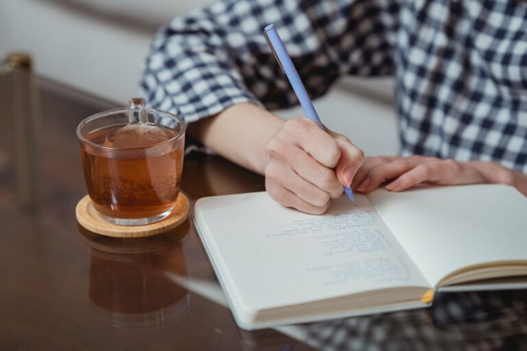 JLNICH blog beats article, Storyteller's Sanctuary: Self-Care Strategies for Writers During NaNoWriMo. Cover image of woman writing in a book with a cup of tea