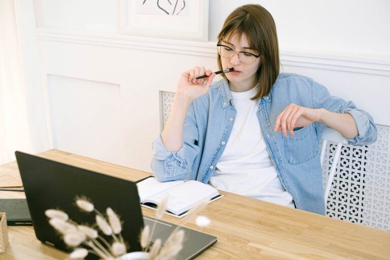 JLNICH blog beats article,Novel-Not-Writing Month: Surviving the Epic Battle Against Writer's Block During NaNoWriMo, cover image of lady sitting and staring at laptop chewing on a pen